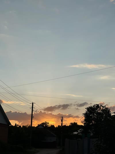 This media file captures a serene sunset scene in Krasnokutsk, Ukraine. The image is oriented vertically, with the sky occupying the left two-thirds of the frame and the lower portion revealing a rural or semi-rural setting.
The sky is a canvas of soft pastels, transitioning from a pale blue at the top to warmer hues of orange and yellow near the horizon. Wispy clouds, tinged with pink and gold by the setting sun, are scattered across the sky. A few power lines crisscross the scene, supported by utility poles, indicating the presence of infrastructure in the area. Silhouetted against the vibrant sky are trees and the dark outlines of houses or other buildings, suggesting a residential or village environment. The bright glow of the sun peeking through the clouds implies it is either just before or just after the sun has dipped below the horizon, typical of a late evening or early morning.
The overall mood is peaceful and tranquil, evoking the quiet beauty of a rural sunset. There are no people visible in the frame. The setting appears to be a common, everyday landscape, rather than a specific event. No text is visible in the image. The weather appears clear with scattered clouds, and the time of day is definitively sunset.