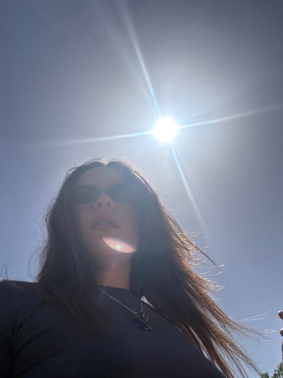 The image is a low-angle shot of a person with long, dark hair and sunglasses, against a bright, clear sky with the sun shining intensely. The person is partially silhouetted and appears to be looking down at the camera. A necklace is visible around their neck. The location, Alcoi/Alcoy, Spain, cannot be verified from the image alone.