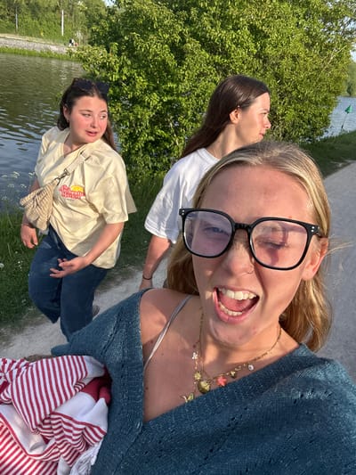 A brightly lit outdoor selfie captures three young women enjoying a sunny day, likely along a river or canal in Marquette-lez-Lille, France. The image is rotated, showing the ground plane on the right.
In the foreground, a blonde-haired woman with black-rimmed glasses pushed onto her forehead grins widely at the camera, her teeth visible. She wears a blue, open-knit cardigan over a light top, adorned with multiple gold necklaces featuring various charms like stars, shells, and a sun. She holds a red and white striped item, possibly a blanket or towel.
Behind her, to the left, another young woman with dark brown hair is seen in profile, looking slightly upwards and away. She wears a white t-shirt, her face illuminated by the bright sunlight.
Further back and to the right, a third young woman with dark hair has sunglasses perched on her head. She wears a light yellow t-shirt with a graphic that reads "Authentic Market CAN SUD" accompanied by a lemon design. She carries a woven, light-colored handbag slung across her body and is dressed in blue jeans. She also looks away from the camera, gazing towards the distance.
The setting features lush green trees and foliage on the left, a calm body of water on the right, and a paved path where the individuals are standing. The clear, bright sunshine indicates good weather, suggesting an afternoon outing. The overall mood conveyed is one of joy and relaxation among friends.