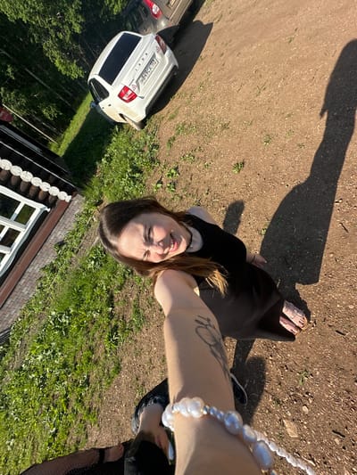 In Bogorodskoe, Russia, a young woman with long brown hair, wearing a black dress and sandals, is captured in a selfie. She is standing on a dirt path with sparse green vegetation. To her left is a wooden house with white-framed windows. A white Lada car is parked on the dirt path behind her. The scene is illuminated by bright sunlight, casting a long shadow of the woman on the ground.