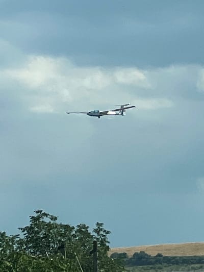 The image shows a biplane in mid-flight against a light blue sky with scattered clouds. The plane is the primary subject, and it appears to be a propeller-driven aircraft, possibly a stunt plane or a crop duster, with its distinctive wing structure visible. The plane is angled with its nose pointing downwards, suggesting it might be performing a maneuver or descending.

In the background, to the right of the plane, there is a sliver of a landscape. It appears to be a field with golden-brown vegetation, likely harvested crops, and a line of green trees. The line of trees could be bordering the field or a road. Some greenery from a tree is also visible in the bottom right corner of the image, suggesting the photograph was taken from ground level.

The lighting suggests it is daytime, likely late morning or afternoon, given the brightness of the sky and the visibility of the landscape. The sky is mostly clear, indicating fair weather conditions. There is no visible text in the image. The overall impression is one of an outdoor scene, possibly in a rural or agricultural area, capturing a moment of flight. The image seems to have been taken from a slight distance, and the plane is not in sharp focus, which might indicate it was moving quickly or the camera settings were not optimized for capturing distant, fast-moving objects.