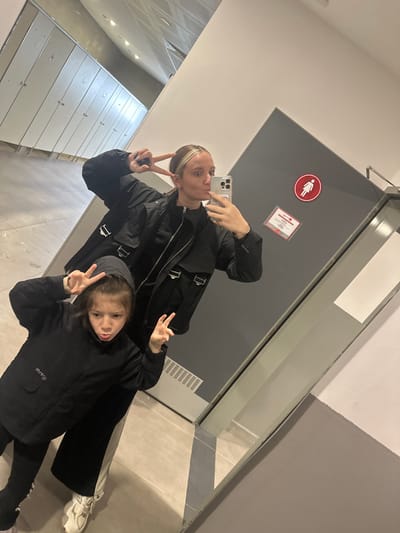 An adult female and a young female child are depicted in a mirror selfie taken indoors in Kemalpaşa, Turkey. The adult, positioned behind the child, has light brown hair with blonde highlights, pulled back, and wears a black jacket. She holds a light-colored smartphone with her right hand, capturing the reflection, while her left hand forms a peace sign. Her lips are pursed. The child, in the foreground, wears a black hooded jacket and dark pants, with both hands forming peace signs. Her expression is direct and solemn.

The reflected environment features a light-colored tiled floor and white or light-grey walls. In the background reflection to the left, a row of eight uniform, light-colored closed doors is visible, suggesting changing rooms or lockers. To the right, a dark grey door is prominent, marked with a red circular sign displaying a white female pictogram, indicating a women's restroom. Below this, a rectangular red-bordered sign is affixed. A white vent runs horizontally near the base of the grey door. The ceiling appears reflective with embedded lighting fixtures.