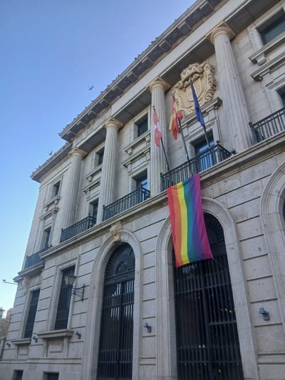 The image shows the facade of a stone building in Ávila, Spain, with a rainbow flag hanging from a balcony. The building is ornate, with classical columns, arched windows, and decorative friezes. Three flags are flying from poles above the balcony: the flag of Spain, another flag with red and yellow stripes, and a blue flag. The rainbow flag, a symbol of LGBTQ+ pride, is prominently displayed below the balcony, partially obscuring a large, dark metal gate. The sky is clear and blue, with a few birds in flight. The perspective is from a low angle, looking up at the building.