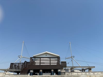 The image depicts the upper portion of a structure resembling a pier or boardwalk, with a small white building on a wooden deck. The building has a pitched roof and horizontal white louvers visible through dark wooden railings. Two tall, slender masts with ladders ascend from the structure, supporting numerous cables that stretch across the bright blue sky. The lower part of the structure is supported by dark pilings. The scene appears to be located outdoors, under a clear sky, possibly in a coastal city. There are no people visible in the image. The overall impression is of a functional, perhaps utilitarian, structure with a clear, open sky as the backdrop. The style and presence of masts and cables could suggest a maritime or communication-related purpose, or simply architectural design elements. The concrete base and weathered appearance suggest it is a permanent fixture. The location is not explicitly identified in the image itself, and there is no visual information within the image that specifies it is Cesenatico, Italy.