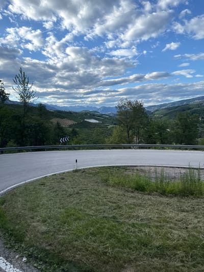 This media file captures a scenic view of a winding road in Spormaggiore, Italy. The road, paved with asphalt and bordered by a white line, curves to the right, flanked by lush green grass on its outer edge. A metal guardrail runs along the left side of the road, separating it from a sloping landscape.
In the distance, the terrain is characterized by rolling hills covered in dense green trees and what appear to be agricultural terraces, possibly vineyards or orchards, suggesting a rural or mountainous region. The backdrop features layers of hazy mountains under a vast blue sky filled with dynamic, streaky white clouds. The lighting suggests it is either early morning or late afternoon, with the sun casting soft light on the scene.
A black and white chevron sign is visible on the left side of the road, indicating a sharp turn ahead. A solitary black post with a white reflective marker stands by the roadside. The overall atmosphere is one of tranquility and natural beauty, inviting a sense of peaceful exploration. There is no visible text in the image, aside from the directional sign.