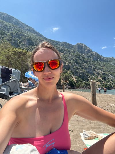 A medium-shot, selfie-style image captures a woman at a beach in Ortaca, Turkey. She is wearing a pink swimsuit and reflective sunglasses. The background includes a mountain covered in dense green vegetation, a beach with a few people on it, and a bright blue sky. The woman is positioned on a wooden pier or deck, with beach sand visible nearby. She is also accompanied by a child.