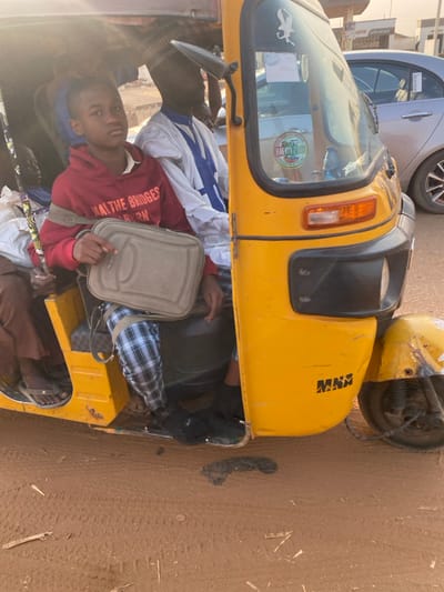 A yellow auto-rickshaw is tightly packed with passengers and luggage. One passenger, wearing a red hoodie, gazes directly at the camera while holding a light-colored bag. Inside the vehicle, plaid and white patterned fabrics mingle with darker bags and more individuals. The auto-rickshaw is parked on a dusty, light brown surface. The vehicle is near a car in the background.