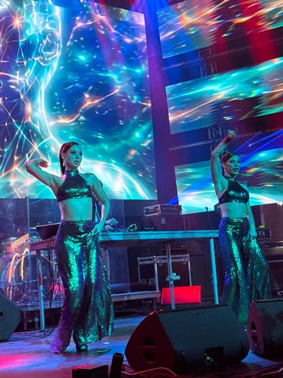 This is a medium shot photograph of two women performing on a stage in what appears to be Misaktsieli, Georgia. The women are dressed in matching, sequined outfits consisting of halter-neck crop tops and wide-legged pants. Both women have their hair pulled back in ponytails. The woman on the left is positioned more towards the front of the stage, with her left arm raised and bent at the elbow, and her right hand resting on her hip. The woman on the right is further back and to the right of the frame, with her right arm extended upwards.
The stage is lit with vibrant, multicolored lights that are projected onto a large backdrop. The backdrop displays a dynamic, abstract visual with flowing lines and colors, primarily blues, greens, and purples, with streaks of orange and yellow. In the midground, there is a DJ booth with turntables and other audio equipment. Various speakers and lighting equipment are visible on the stage and in the foreground. The overall atmosphere is one of a live musical performance or dance show.