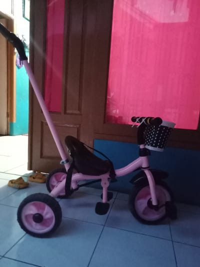 The scene, located indoors in Ciputri, Indonesia, presents a child's pink tricycle positioned slightly right of center, facing right, on a light blue tiled floor with dark grout lines. The tricycle features a pink frame, pink wheel spokes, black tires, a black seat with an attached strap, black pedals, and black handlebars adorned with a white basket detailed with black polka dots. A long, pink push handle extends upwards and to the left from the rear of the tricycle, culminating in a black grip. To the left, an open light-brown wooden door frame reveals a room with light blue walls and a lighter floor. Two yellow, closed-toe sandals are placed on the tiled floor near this doorway. To the right, a vertical wooden door panel stands, partially obscured by a large, uniformly red curtain or blind. A section of a light blue wall is visible below the red curtain and to the right of the wooden door. No individuals are present within the frame.