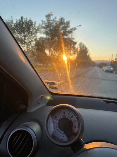 The image depicts the interior of a car, looking through the windshield at sunset. The sun is low on the horizon, casting a warm orange glow and creating lens flare. Trees and a road are visible outside, with a few cars in the distance. The car's dashboard is in the foreground, showing a circular tachometer with a needle pointing between 0 and 1. An air vent is also visible to the left. The scene is set in Delchevo, North Macedonia, evident by the light pollution and mountainous horizon typical of the region.