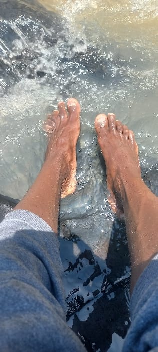 The image displays a first-person perspective of an individual's bare feet and lower legs submerged in flowing water, likely a river or stream, located in Kipkenyo ward, Kenya. The individual is wearing grey shorts or trousers, visible from mid-thigh down. Both feet, exhibiting a dark skin tone, are positioned with toes pointed away from the viewer, partially immersed in clear to slightly murky water. Sunlight reflects off the water's surface, creating bright glints and illuminating small water droplets on the skin of the legs and feet. The water shows ripples, splashes, and small pockets of foam, indicating movement. Dark, possibly rocky or muddy riverbed material is discernible beneath the water, particularly between the legs and in the background. No other individuals or distinct man-made objects are visible, suggesting a natural outdoor environment.