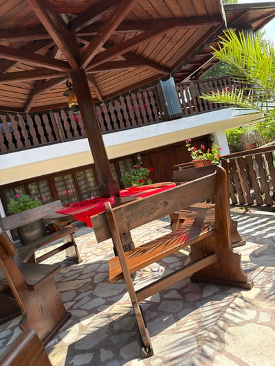This image depicts an outdoor seating area within the city of Zlati Voyvoda, Bulgaria. The central feature is a dark brown octagonal wooden pergola, supported by a thick central post from which a decorative lantern hangs. Below the pergola, multiple rustic dark brown wooden picnic-style tables and benches are arranged on a patio. Several tables are draped with red fabric runners, some exhibiting woven patterns.
In the background, a two-story building with a white base and a cream-colored facade on the lower level is visible. This level features multiple dark wooden-framed windows, some with lace curtains, and window boxes containing potted pink flowering plants, likely geraniums. The upper story of the building showcases a wooden balcony with a dark brown, intricately carved or turned balustrade. Above the balcony, a dark wooden roof structure with scalloped eaves extends.
Green vegetation, including a prominent vibrant green palm frond in the upper right quadrant and other foliage in the background, borders the scene. The ground is paved with irregularly shaped flagstones in various shades of grey and beige, interspersed with dark grout. The area is brightly lit by direct sunlight, casting strong, distinct shadows from the furniture and structures onto the paved surface. No individuals are present in the image.
