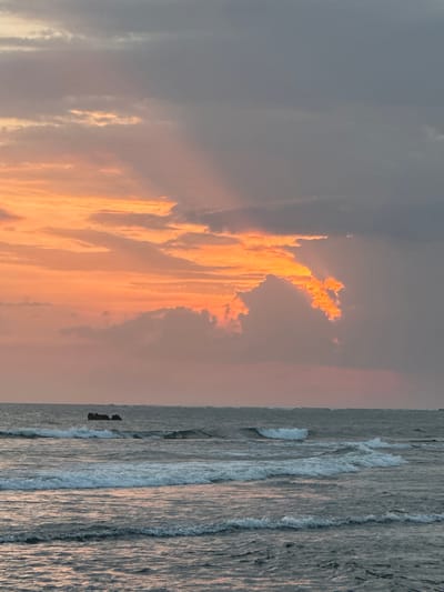 The image shows a dramatic sunset over the ocean in North Kuta, Indonesia. The sky is filled with an array of colors, from fiery orange and pink near the horizon to a deep gray in the upper portions. Rays of light pierce through the clouds, creating a dynamic and ethereal effect. In the distance, a rocky outcrop or small island juts out of the choppy water. The ocean itself is a mix of gray and blue, with white waves breaking closer to the shore, indicating a moderate swell. The overall scene is one of natural beauty and a powerful display of atmospheric conditions.