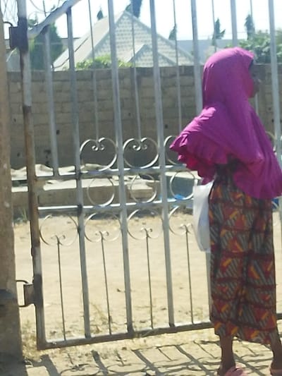 An outdoor scene in Darmanawa, Nigeria, depicts an individual standing partially obscured behind a light-colored metal gate. The person, likely female, is dressed in a vibrant pink head covering that drapes over her shoulders and a long, patterned skirt featuring geometric designs in red, orange, yellow, and grey. Pink sandals are visible on her feet. She holds a white plastic bag in her left hand. The metal gate comprises vertical bars with decorative heart-shaped and circular elements. Behind the gate, a low concrete block wall is present, with several houses featuring patterned sloped roofs and distant green foliage visible under a clear, bright sky. The ground in the foreground and through the gate is sandy or dusty, and shadows are cast by the gate due to direct sunlight.