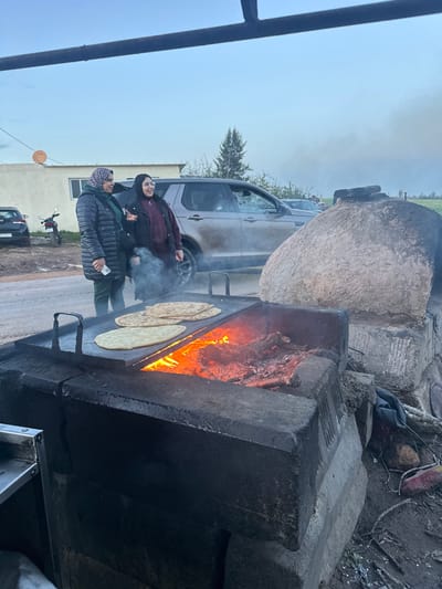 The image captures an outdoor cooking scene with two women observing a traditional fire-powered setup. The main focus is a large, rectangular cooking station constructed from dark, robust stone or concrete, situated centrally in the frame. Inside this structure, a vibrant fire blazes with bright orange and red flames, fueled by wood or charcoal, and producing a significant amount of smoke that drifts upwards and to the left. On a flat griddle-like surface positioned above the fire, several pieces of round or oval flatbread are cooking. Some appear light and freshly placed, while others show browning from the heat.

To the left of the cooking station, two women are present. The woman in the foreground wears a dark puffer jacket, green trousers, and a patterned knit head covering. She is holding a white smartphone in her left hand and looking towards the cooking area. Behind her, another woman is dressed in a dark maroon or deep purple jacket, dark trousers, and a dark head covering (possibly a hijab). She is smiling and looking towards the fire and flatbreads. Both women are standing on what appears to be a dirt or gravel surface.

Behind the cooking station, a large, light-colored, textured, and somewhat irregular spherical or ovular object, possibly a traditional clay oven (like a tannur or tabun) or a large pot, is visible. Further in the background, a light-colored SUV is parked, partially obscured by the cooking setup and the women. Beyond the SUV, a tall, dark green coniferous tree stands out against the sky. A paved or unpaved road is visible further back, with a smaller dark car and a motorcycle parked alongside it. Green fields stretch into the distance beyond the road.

The sky is overcast, appearing light gray or white, suggesting either early morning, late afternoon, or simply a cloudy day. The overall atmosphere appears casual and domestic, centered around the traditional food preparation. No discernible text is visible in the image.