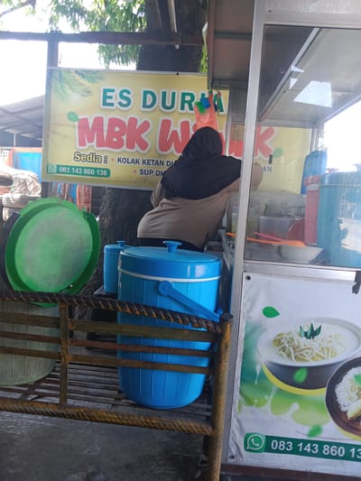 The image depicts an outdoor food stall in Sarimulyo, Indonesia, illuminated by daylight. A person, viewed from behind, is positioned at the stall counter, wearing a black head covering (hijab) and a beige-colored long-sleeved top.
The upper section of the stall displays a yellow banner with green and red text. Prominently featured are "ES DURI MBK W" (likely "Es Durian Mbak W") and "Sedia:" (Available:), followed by "KOLAK KETAN DU" (Kolak Ketan Durian) and "SUP DU" (Sup Durian). A green WhatsApp icon with the phone number "083 143 860 136" is printed on the lower left of this banner. Stylized palm fronds are illustrated in the upper left corner of the yellow banner.
The stall features a glass display case, reflecting bright light and structures from the environment. Inside the case, multiple clear plastic containers, white bowls, and orange and pink serving utensils are visible. To the right, behind the glass, a tall, narrow blue container and a pink container are discernible.
Below the stall counter, a lower white banner or sign features an image of two dessert bowls. One bowl contains a white, creamy substance topped with shredded white ingredients (resembling cheese or coconut) and a green leaf garnish. A second, partially visible bowl to the right contains a similar white base with textured white and green toppings. This lower banner also includes a green WhatsApp icon and the phone number "083 143 860 13".
In the foreground, a rusty, dark brown, twisted metal railing or frame runs horizontally. Behind this railing, two large blue insulated plastic containers, one with a blue carrying handle, rest on a metal grate or shelf. To the left of these containers, a large green plastic lid or shallow basin leans upright.
The background behind the stall shows a tree trunk and green foliage. The ground surface appears to be paved with concrete.