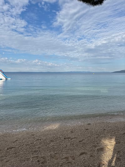 The image depicts a coastal scene in Makarska, Croatia. In the foreground, a pebble beach is visible, with small waves gently lapping at the shore. The water is clear and turquoise, transitioning to a deeper blue further out. In the distance, a long, thin buoy line stretches across the water, indicating a boundary or swimming area. To the left, an inflatable water slide is partially visible, protruding from the water. The horizon is marked by a hazy, mountainous coastline. The sky above is partly cloudy, with dramatic white clouds against a blue backdrop. A dark silhouette of pine needles is visible in the upper right corner, suggesting vegetation overhead. The lighting suggests it is daytime, possibly late morning or early afternoon, with a slight shadow cast across the beach. No people are visible in the image.