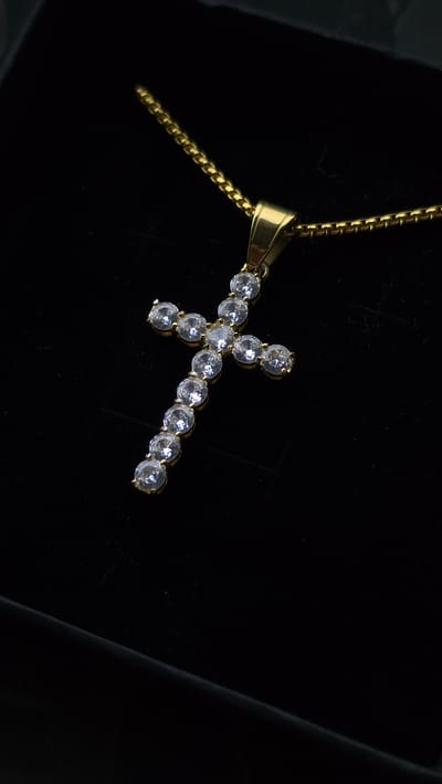 A gold chain necklace with a cross pendant is displayed on a black surface. The cross pendant is adorned with clear, round gemstones. The background is dark, with subtle lighting creating highlights on the jewelry. The image does not contain any information about the city of Liliak, Bulgaria, or any people.