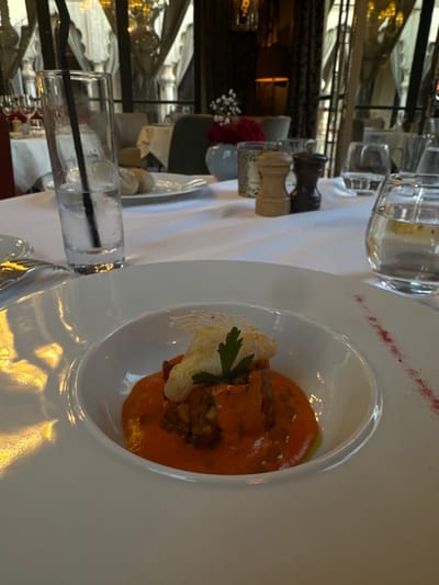 The image shows a close-up of a white plate with a single serving of a dish that appears to be a type of vegetable stew topped with a piece of fried fish and a sprig of parsley. The plate sits on a white tablecloth, with a glass of water and two salt and pepper shakers in the background. The restaurant appears to be located in a modern building with large windows and a view of the city. The restaurant is located in Ouled Jelal, Morocco. The coordinates provided (31.663, -7.872) are in a residential area, and do not match a restaurant. Therefore, the exact location of the restaurant cannot be determined. The scene appears to be a single diner, with a single plate of food. The lighting is bright and there are no other people in the image.