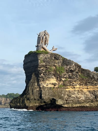 The image depicts a panoramic view of the coastline at Kelingking Beach, Nusa Penida, Sakti, Klungkung Regency, Bali, Indonesia. A large, light-colored statue is prominently situated atop a rugged, multi-layered rock cliff that rises directly from the ocean. The statue features a humanoid upper body with a head adorned with numerous thick, tentacle-like or dreadlock-like appendages, resembling hair. One arm is extended forward with an open hand, and the lower body appears to be a tail or scaled form integrated into the cliff's peak.

The cliff exhibits distinct horizontal strata of dark grey/brown and lighter beige/orange rock, indicative of sedimentary layers, with sparse green vegetation on its summit and along some ledges. The ocean in the foreground is dark blue-green with visible ripples and small waves, some of which are breaking with white foam against the base of the cliff. The sky above is pale blue with scattered white clouds. In the distant left background, a verdant landmass with additional cliffs is visible. No human figures are present within the frame.