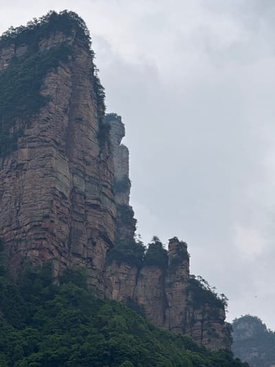The image depicts a mountain landscape in Zhonghu, China. The mountains are characterized by steep, layered rock formations. Green vegetation covers parts of the mountains, particularly on the right side of the frame. The sky is overcast with grey clouds.