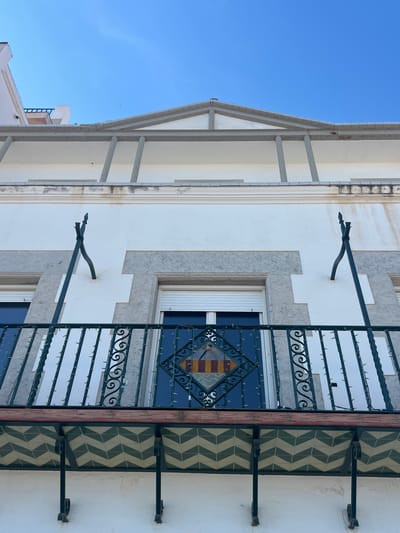 The image depicts a section of a building facade in Sant Pol De Mar, Spain. The architecture features a white exterior with grey stone accents. A window with white blinds is visible, partially obscured by an ornate, dark-colored wrought-iron balcony. The balcony has a decorative central element that includes yellow and red hues. To the right of the balcony, there are tiled panels with a chevron pattern in shades of green and white. Supporting the overhanging roof structure on the left, there are vertical white pillars. The sky above is clear and blue. Extending from the wall, there are dark-colored, curved metal supports, which give the impression of adding support for a canopy or sunshade.