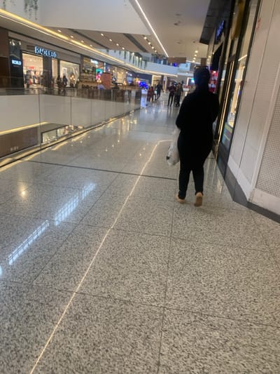The image displays an interior view of a multi-level shopping mall, specifically identified as Salvador Norte Shopping in Lauro de Freitas, Brazil. In the foreground, a person viewed from behind, dressed in dark clothing (black top and pants) and light-colored flat shoes, walks away from the camera on a highly polished, speckled gray and white tiled floor. This individual carries a white plastic shopping bag in their left hand.
To the left of the walking individual, a transparent glass balustrade runs parallel to the path, separating the current level from a lower mall floor which is partially visible. Reflections of the ceiling and distant mall elements are evident on the balustrade. To the right, a dark-colored store facade with a large display window features vertical, ribbed panels to its right.
In the mid-ground, further down the corridor, various store fronts are visible, including a prominent "Riachuelo" sign on a department store. Several other indistinct figures of people are present in the background, walking or standing within the mall environment. The mall ceiling features linear strip lighting, which casts distinct reflections on the polished floor, along with some suspended green foliage elements. The overall scene is well-lit by artificial illumination.