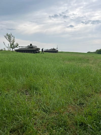 The image depicts two military tanks positioned in a grassy field. A single tree is visible near the front of the first tank, on the left side of the image. A person in military attire is standing between the two tanks. The sky is overcast with numerous clouds. The location is Kostena Reka, Bulgaria.