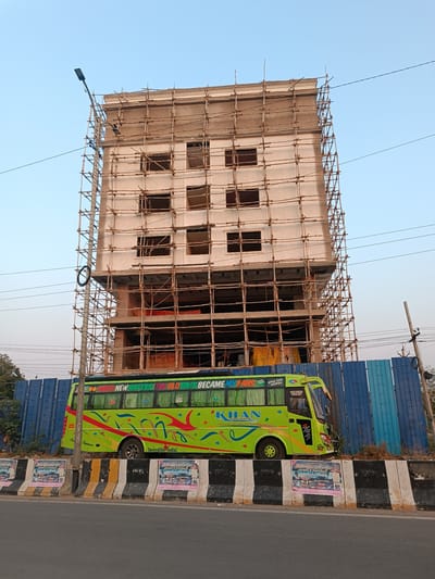 The image depicts an urban scene in Anantapur, India, featuring a multi-story building under construction and a parked bus.

**Building:** A six-story concrete structure, light beige in color, is visible directly behind a road. The building is heavily covered with bamboo scaffolding on all observable sides. Numerous rectangular window openings are present across the top five visible floors, lacking glass or frames. The ground floor and first floor show partially completed concrete work and a large open frontage.

**Bus:** A lime-green coach bus is parked on the right side of the road, parallel to the construction site. The bus is adorned with red, blue, and green stylized patterns. Text on its side reads "I DON'T NEED NEW HATERS. THE OLD ONES BECAME MY FANS." and "KHAN HOLIDAYS GROUP OF CONCERN". Additional text includes "APNA TIME AAYEGA" and "Incredible India!". The rear license plate, "AP39 TB 1111", is visible, confirming registration in Andhra Pradesh.

**Road and Median:** In the foreground is an asphalt road. A concrete median barrier, painted with alternating black and white stripes, runs along the edge of the road, separating it from the bus and construction site. Several rectangular posters, depicting local advertisements or events, are affixed to the median barrier.

**Environment:** A tall, slender metal light pole with two attached streetlights stands on the left, slightly in front of the building. Multiple utility wires traverse the frame horizontally. To the right of the bus, a tall blue corrugated metal construction fence obscures the lower section of the construction area. Sparse, dry vegetation is visible behind this fence. The sky above is clear and light blue. No individuals are clearly identifiable in the immediate foreground.