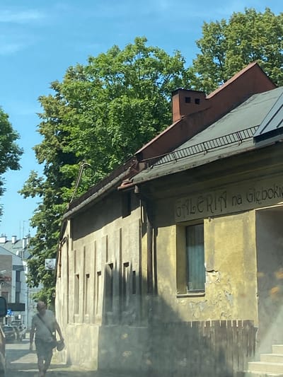 The image shows a street scene in Bielsko-Biała, Poland. A man wearing a light-colored t-shirt and shorts walks on the sidewalk. He carries a bag over his shoulder and holds a circular object in his hand. The man is walking in front of a weathered, yellow building with signage that reads "GALERIA na Głębokiej". A wooden fence runs along the front of the building. Trees with green foliage line the street. In the background, other buildings and parked cars are visible. The sky is blue with some clouds.
