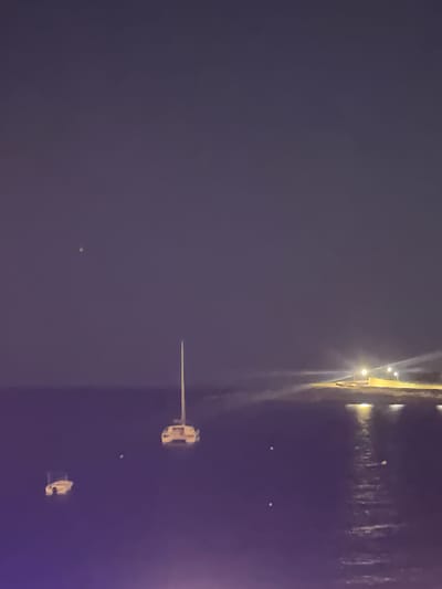 The image depicts a nighttime scene in Saint Paul's Bay, Malta, with a focus on the bay itself. Several small boats are visible on the water. Along the shoreline, lights from buildings and streetlights illuminate the edge of the bay. The overall impression is one of a calm, dark night with faint lighting along the coast.