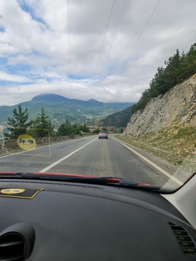 The image is a point-of-view shot from inside a red car, looking out at a winding mountain road in Artvin, Turkey. The road is asphalt with a white center line and a lighter white line marking the shoulder. On the right side of the road, there is a steep, rocky embankment covered with scattered green vegetation and pine trees. On the left side of the road, there is a concrete barrier, followed by a drop-off with trees and a distant view of a valley with buildings and a large, mist-covered mountain range in the background. Power lines are visible stretching across the sky, supported by poles. A silver car is driving ahead on the road. The sky is cloudy with patches of blue. A yellow sticker with a smiley face wearing sunglasses is affixed to the car's dashboard on the left side.