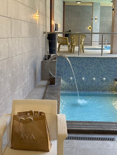 The image depicts a resort area, likely a spa or hotel, with a small indoor pool. The pool is tiled in blue and white squares and features a small waterfall cascading into it from a water feature. Several small water jets are visible on the tiled wall behind the pool.
In the foreground, a light-colored wicker chair sits empty, with a brown canvas tote bag resting on it. The bag has the logo "AYĀ RESORT" printed in white. Behind the chair, a metal railing separates the seating area from the pool.
Further back, two empty yellow plastic chairs are placed on a tiled patio. The walls are covered in large, light gray tiles with a subtle veining pattern. Several wall-mounted lights with warm illumination are visible on the walls, casting a soft glow. In the background, glimpses of additional pool areas and architectural elements are visible. The overall impression is one of a clean, modern, and relaxing resort environment. The image does not contain any people. The location is stated to be Ayn al Basha, Jordan.