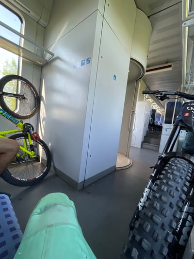 The image depicts the interior of a train carriage, likely in Merichleri, Bulgaria, designed to accommodate bicycles. Two bicycles are visible, one a bright yellow mountain bike mounted vertically on the left, and another black mountain bike in the foreground, its front wheel and handlebars prominent. The train carriage features light-colored walls with visible signage indicating "2. KL." (2nd class) and a "WC" symbol. The flooring is grey, and seats are visible in the background, arranged in rows. A circular door mechanism is also noticeable. A portion of a person's leg is visible on the left, and another person's knee covered by a mint green fabric is in the lower right corner. Sunlight streams in from a window on the left, casting shadows.