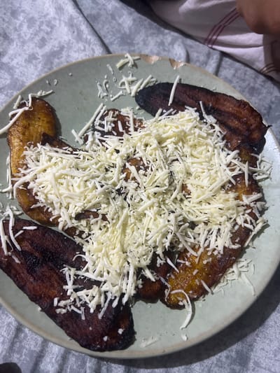 A plate of fried plantains, also known as maduros, topped with shredded white cheese sits on a grey, textured surface that appears to be a tablecloth. The plantains are dark brown and appear to be ripe, with some slightly charred edges. The cheese is finely shredded and generously scattered over the top of the plantains. The plate is a light green, ceramic dish. The overall lighting suggests an indoor setting, possibly in a home environment in Villa de Cura, Venezuela.