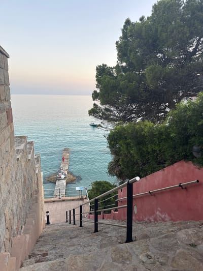 The image depicts a scenic view of the sea from a stone staircase. The staircase itself is a prominent feature, constructed from large, rough-hewn stones. A red wall runs alongside the staircase, with a metal railing providing a safety barrier. Lush green trees overhang from the top left of the frame, casting shadows.

Below the staircase, the turquoise sea is visible. A small concrete jetty extends into the water, with a few people gathered on it. One person in a red top is standing, while others are seated. A small boat is moored near the jetty. The water is calm, with gentle ripples on the surface. There are also a couple of yellow buoys floating in the sea.

In the distance, the horizon is visible, with the sky appearing pale and hazy, suggesting either early morning or late afternoon. A faint rainbow-like arc is visible in the sky above the sea. The overall mood is tranquil and serene.

The location context is Es Camp De Mar, Spain. There is no visible text in the image.