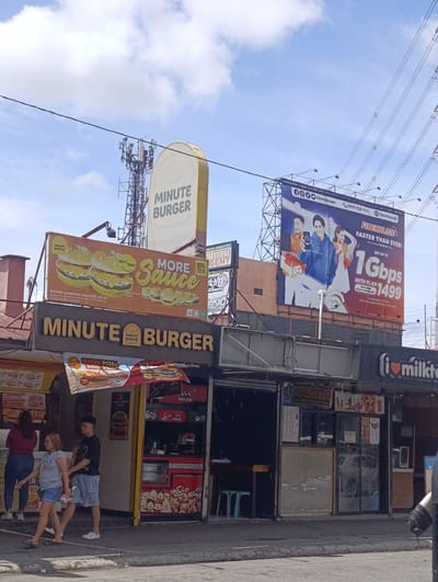 This image captures a street scene in Imus, Philippines, under a bright, partly cloudy sky, suggesting daytime. The primary focus is on a row of small food stalls and signage.

A prominent "MINUTE BURGER" sign dominates the upper portion, with another larger "MINUTE BURGER" sign below it, featuring images of burgers and advertising "MORE Sauce" and a "BUY 1 TAKE 1" promotion. To the right, a large billboard advertises "FIBERBLAZE" internet service, highlighting speeds up to 1 Gbps for plan 1499.

In the foreground, several people are visible. Two women walk along the sidewalk, one in a red top and jeans, the other in a grey t-shirt and denim shorts. A young man in a black t-shirt and blue shorts walks behind them. Behind the Minute Burger stall, more people are seen, and behind them, a stall labeled "Sio Ai House" is visible, showcasing fried chicken. Another stall displays "PCSOLOTTO."

The scene depicts a bustling commercial area with various food vendors and services. The stalls are open, suggesting they are actively serving customers. The overall impression is one of a typical urban street in the Philippines during the day, characterized by vibrant signage and pedestrian activity.