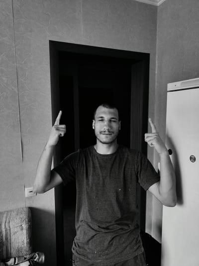 In a black and white indoor photograph, a young man stands in a doorway, looking towards the camera with a slight smile and a wink. He is wearing a dark t-shirt and shorts, and his arms are raised with index fingers pointing upwards. The background features textured wallpaper on the left and a white appliance, possibly a refrigerator, on the right. A dark doorway is visible behind him, leading into a dimly lit interior. A glimpse of a patterned chair and a light switch can be seen to the left of the frame. The image appears to have been taken indoors, possibly in a residential setting, with natural or ambient lighting. No specific time of day or weather conditions are discernible. A circular sticker with text is visible on the white appliance on the right, but the text is too small to be read clearly. The overall mood of the image is casual and playful, conveyed by the man's gesture and facial expression.