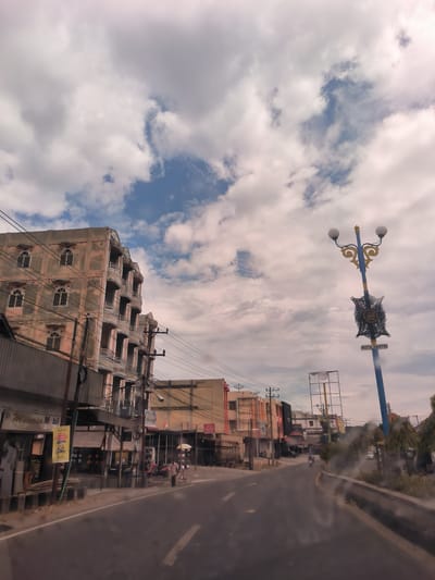 This street-level photograph captures an urban scene in Bireuenmeunasahcapa, Indonesia, under a sky filled with a mix of large white and grey clouds, with occasional glimpses of blue. The time of day is daytime, with diffused light.

On the left, a multi-story building with a weathered, possibly pink-hued facade, features multiple arched windows and balconies. Dense utility poles and numerous overhead wires crisscross in front of and around this building. Below, commercial activity is evident with various storefronts. A yellow sign is prominent, displaying the "maxim" logo and the text "Pesan perjalanan" (Order a trip). Another partially visible sign near the building reads "Mobil" (Car).

The asphalt road, possibly viewed from inside a vehicle due to reflections or smudges in the foreground, has a white dividing line. Further down the street, a few people are visible on the sidewalk; one individual is holding an umbrella. A motorcycle is also seen on the road in the distance.

On the right side of the image, a distinctive blue and gold street light or decorative pole stands tall. It features two spherical lamps at the top and an ornate black metal plaque affixed below the lights. This plaque has text in Arabic script, and below it, a sign clearly states "KOTA SANTRI" (City of Santri). The scene depicts everyday life on a bustling street, characterized by a mix of residential and commercial buildings, urban infrastructure, and local activity.