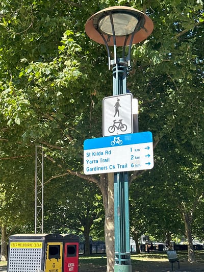 A sign post displays a pedestrian and bicycle symbol along with trail directions. St Kilda Rd is 1 km, Yarra Trail is 2 km, and Gardiners Ck Trail is 6 km in the direction indicated by arrows. Below the sign, a trash receptacle with separate compartments for recycling and general waste reads "KEEP MELBOURNE CLEAN." A vintage-style streetlight with a circular reflector is mounted at the top of the pole. The scene is framed by leafy green trees.