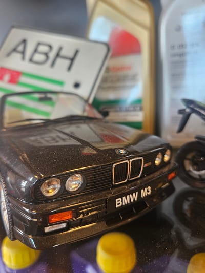 This is a close-up, eye-level, shallow depth of field shot of a black BMW M3 toy car. The car is dusty and positioned on a dark surface with two out-of-focus yellow caps in the foreground. Behind the car, partially visible, are several bottles, including one with a prominent red circular label and green stripes, and another clear bottle with text. A white sign with the letters "ABH" in black lettering and green and red stripes is also visible in the background, as is a blurred motorcycle. The scene appears to be indoors, with the lighting suggesting a controlled environment, possibly a display or collection. There are no people present. The location in Bemmel, Netherlands, is not discernible from the image itself.