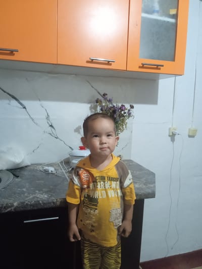 In a domestic kitchen, a young child stands between a countertop and cabinets. The child, wearing a yellow t-shirt with graphics, is facing the viewer. Behind the child, a bouquet of dried flowers is arranged on a countertop next to a white pot. The kitchen features marble-patterned backsplashes and orange overhead cabinets. To the right of the child, electrical outlets are visible on a white wall. The lower cabinets are dark brown. The scene appears to be in a residential setting within Амангелді ауылдық округі, Kazakhstan.