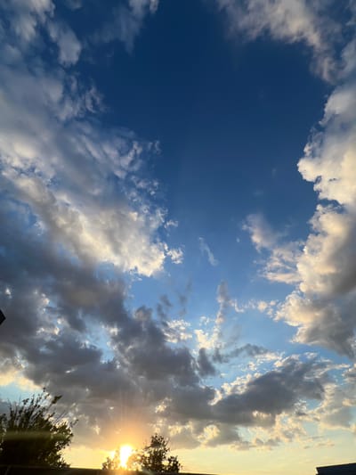 The image captures a sunset over a rural landscape, likely in Ленинское сельское поселение, Ukraine. The sky is a vibrant mix of deep blue and lighter shades, populated by scattered cumulus clouds. The sun, a bright, glowing orb, is partially obscured by trees and low-lying structures at the horizon, casting warm, golden rays and creating a lens flare effect. Silhouetted trees and branches are visible in the foreground, framing the lower part of the scene. The overall impression is one of a serene and picturesque end to the day.
