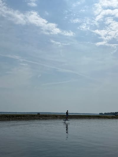 The image depicts an outdoor, daylight scene featuring a single individual on a body of water under a partially cloudy sky, located in Montgomery, United States. A male figure stands in the midground, silhouetted, upright on a hydrofoil board, holding a paddle vertically. His reflection is visible in the calm, dark blue water. Behind the individual, a narrow strip of land, likely a sandbar or shallow bank, stretches horizontally across the frame. A light-colored vehicle, possibly an SUV, is visible on this land strip to the left of the frame's center. Beyond the land strip, more water extends to a distant, tree-lined shoreline on the horizon. The sky above is light blue with scattered white clouds and faint linear cloud formations.