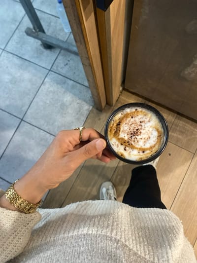 An overhead, downward-angled shot captures a person's upper torso, right arm, and lower legs in an indoor setting within Monastir, Tunisia. The individual holds a black mug filled with a coffee beverage featuring frothed milk, decorative latte art or syrup swirls, and dark sprinkles on its surface. The person wears a light-colored, textured knit sweater, black trousers, and light-toned athletic shoes on the visible right foot. A gold-toned link bracelet adorns the left wrist, and a gold-toned signet ring with a light-colored face is on the right ring finger. The floor exhibits two distinct sections: grey square tiles with dark grout lines on the left, and lighter brown wood-look plank flooring on the right. In the background, a wooden door frame or paneling is visible on the right, while a metal fixture and a clear plastic bottle are present on the left.