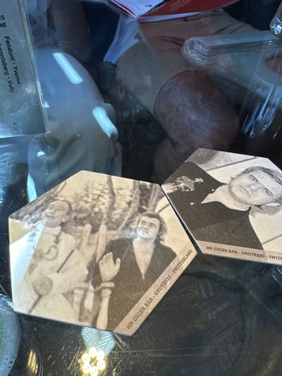 The image presents a close-up, angled view of two hexagonal coasters resting on a reflective, possibly glass, surface, likely within the H.R. Giger Bar in Gruyères, Switzerland, near Epagny.

The top coaster features a black and white portrait of artist H.R. Giger, depicted with an Oscar statuette in front of him. The text "HR GIGER BAR - GRUYERES - SWITZERLAND" runs along its upper right edge. The bottom coaster, partially visible and overlapping the top one, also displays a black and white, intricate, and surreal artwork typical of Giger's style, with the same "HR GIGER BAR - GRUYERES - SWITZERLAND" text along its edge.

The reflective surface heavily mirrors the surroundings, showing indistinct shapes that suggest a person's limb (perhaps an arm or leg), portions of light-colored clothing or objects, and a distinctive circular light fixture composed of multiple small, bright bulbs in the lower right.

In the bottom-left corner, a section of a menu or price list is visible through the reflective surface. It displays text such as "Fendant · Yvorne," "hannisberg · Vully," and a price of "5.50," indicating typical offerings in a Swiss bar or cafe. The scene captures a moment in a unique, themed establishment, highlighting its artistic ambiance through its custom coasters.
