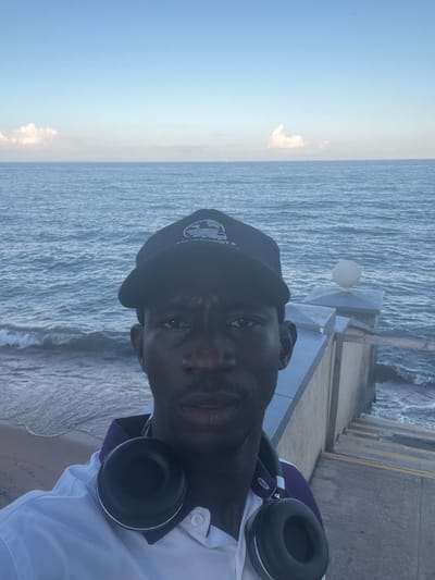 A medium-shot, slightly tilted, head-and-shoulders portrait of a Black man with headphones around his neck and a black cap on his head, positioned on a boardwalk with the ocean as the backdrop. The man is the primary subject, taking up a significant portion of the frame. He is wearing a white collared shirt. The ocean waves are visible on the right of the frame. The sky is light blue and clear on the left. The boardwalk is gray concrete. A rounded lamp post is to the right of the man's head. The precise location in the city of Dowh, Malawi is not deducible from the image alone.