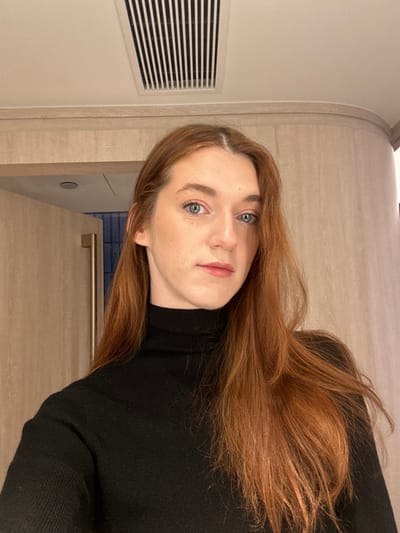 A fair-skinned woman with auburn hair and light eyes poses for a portrait. She wears a black turtleneck sweater. The background appears to be an interior space with beige walls and a vent with horizontal slats on the left. A portion of a cabinet or countertop is visible, suggesting the setting might be a kitchen or bathroom. The woman's face is angled slightly towards the camera, displaying a subtle, neutral expression. The photo appears to be taken in portrait mode.