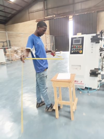 A man is standing in a large, industrial-type building with a high ceiling, possibly in Akute, Nigeria. He is measuring the distance between a wooden structure and a large white machine with a yellow measuring tape. The machine features a control panel with various buttons and displays. The wooden structure appears to be a makeshift table or platform. The floor is a light, reflective color.