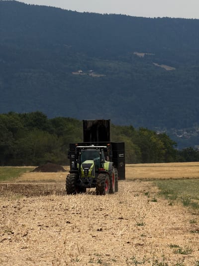 A tractor with a trailer is operating in a field near Reignier-Ésery, France. The tractor is green and black and appears to be hauling material in the black trailer. The field is dry and stubbly, suggesting it may have recently been harvested. In the background, there are trees and a large hill covered in dark green trees. The sky is overcast. The scene appears to be a rural agricultural setting.